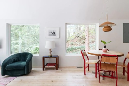 Cozy interior featuring a green chair, wooden table with red chairs, lamp, and large windows overlooking greenery.
