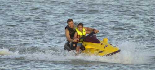 A man and a child ride a yellow jet ski on a calm body of water, both smiling and enjoying the ride.