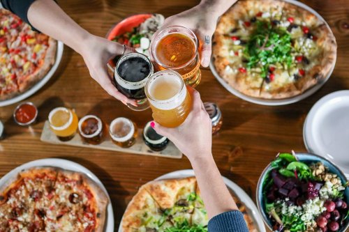 Hands raising glasses of beer over a table with various pizzas and a salad, celebrating with food and drinks.