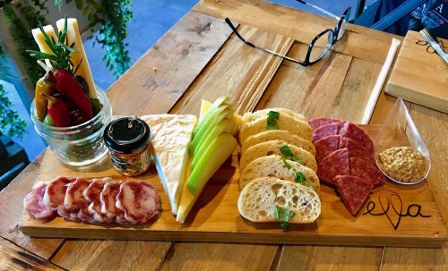 A wooden platter with assorted cheeses, cured meats, bread, mustard, and pickled vegetables, set on a rustic table.