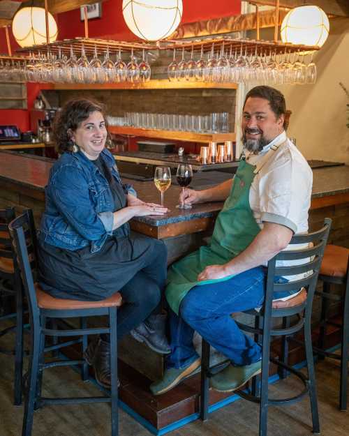 A woman and a man sit at a bar, smiling, with drinks in hand and glasses hanging above them. Cozy restaurant setting.