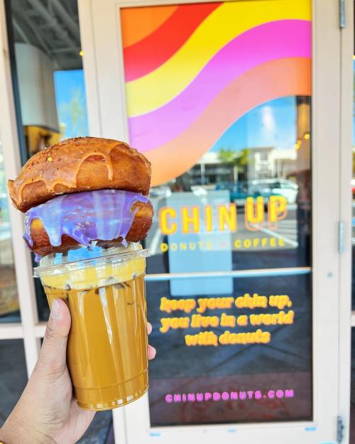 A hand holding a coffee drink and a donut sandwich in front of a colorful donut shop window.