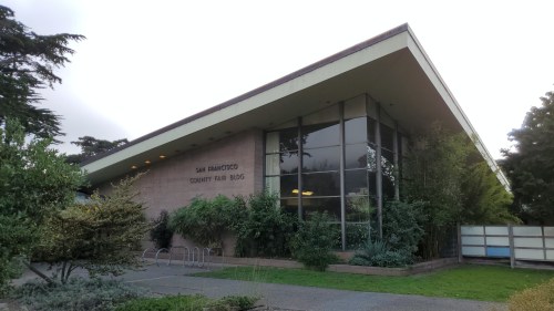 Modern building with large windows, surrounded by greenery, featuring a sloped roof and "San Francisco Community Fair Bldg" sign.