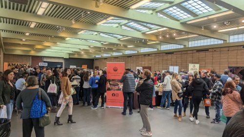 A bustling indoor market with many people browsing booths and enjoying food in a spacious, well-lit venue.