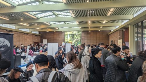 A crowded indoor event with people mingling, booths set up, and large windows letting in natural light.