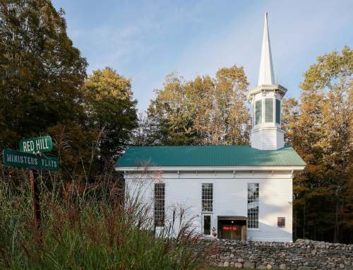 A white church with a green roof and steeple, surrounded by trees, next to a sign for Red Hill and Ministers Flats.