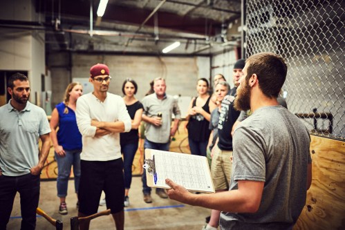 A group of people attentively listens to a speaker holding a clipboard in an industrial setting.