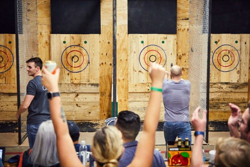 Two men throw axes at wooden targets while a cheering crowd watches in a lively indoor venue.