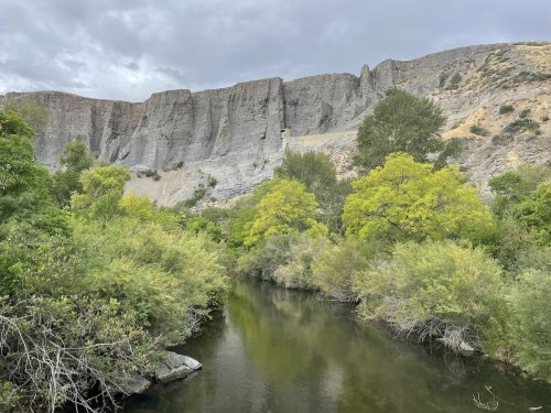 A serene river flows through lush greenery, with towering cliffs in the background under a cloudy sky.