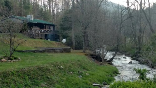 A cozy cabin by a river, surrounded by bare trees and grassy land, with mountains in the background.