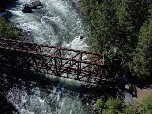 Aerial view of a rusted metal bridge over a rushing river, surrounded by lush green trees and rocky banks.