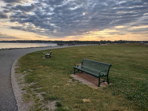 A peaceful park scene with two green benches facing a calm waterway under a cloudy sunset sky.