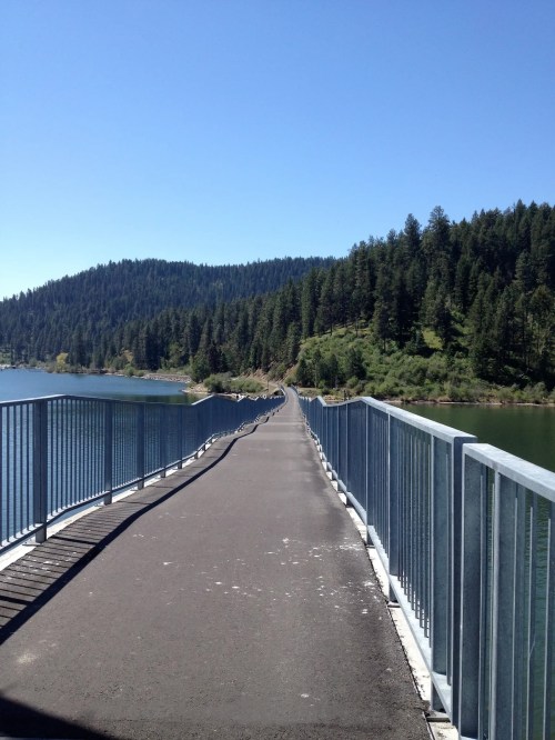 A long pathway stretches over a calm lake, surrounded by lush green hills under a clear blue sky.
