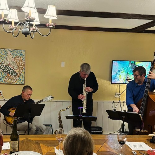 A small jazz band performs indoors, featuring a guitarist, saxophonist, and bassist, with a wine bottle on the table.