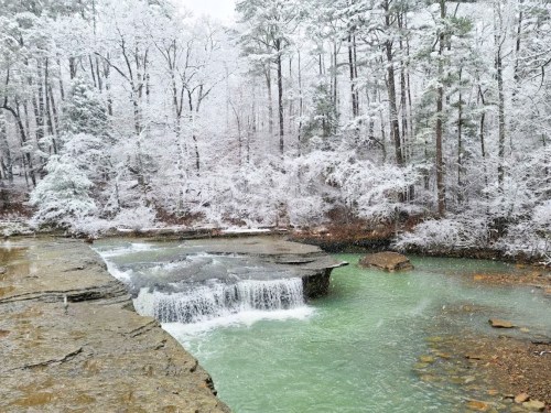 A serene winter scene featuring a snowy landscape, a waterfall, and a turquoise pool surrounded by trees.
