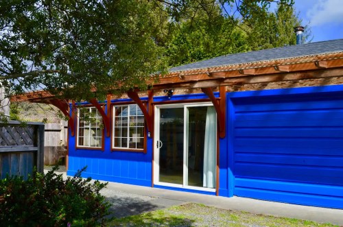 A bright blue house with wooden accents and large windows, surrounded by greenery and a gravel path.