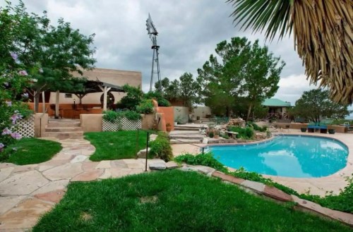 A serene backyard with a pool, stone pathways, lush greenery, and a windmill in the background under a cloudy sky.