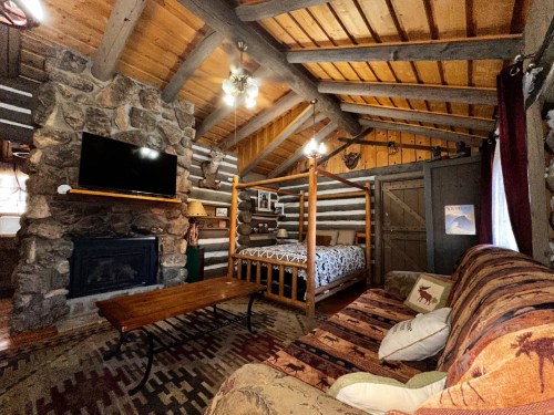 Cozy cabin interior featuring a stone fireplace, wooden beams, a bed, and a comfortable sofa with rustic decor.