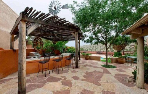 A scenic outdoor patio with a wooden pergola, bar stools, and a windmill in a lush garden setting.
