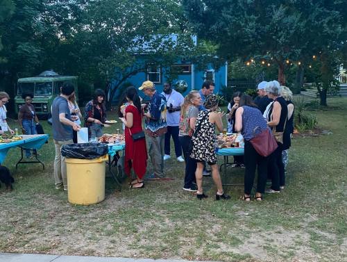 A diverse group of people gathered around tables with food in a grassy outdoor setting during a social event.