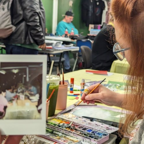 A person holds a photo while painting at a table filled with art supplies in a creative workspace.