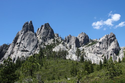 Majestic rocky peaks rise above a lush green forest under a clear blue sky with a few scattered clouds.