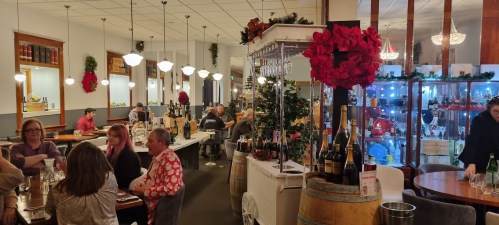 A festive restaurant interior with holiday decorations, tables filled with diners, and a wine display in the background.