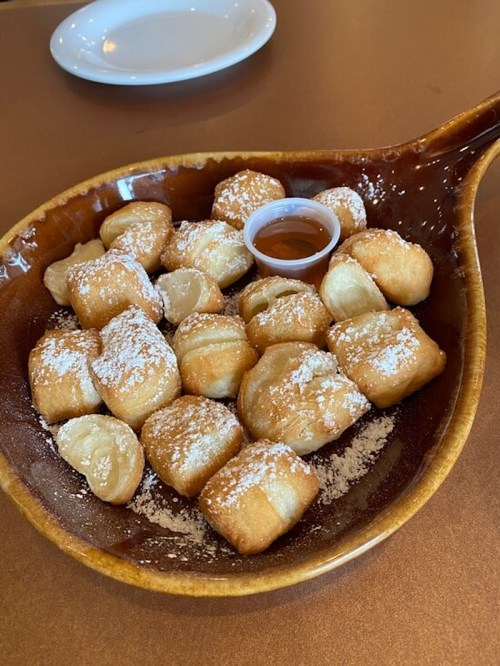 A wooden bowl filled with golden, powdered sugar-dusted dough bites and a small cup of syrup.