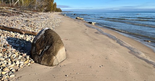 A sandy beach with smooth stones and a large rock, bordered by calm water and trees in the background.