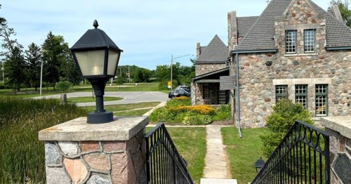 A stone house with a gray roof, surrounded by greenery and flowers, viewed from a pathway with a lamp post.