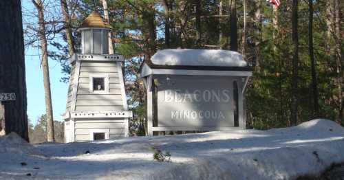 Sign for "Beacons Minocqua" next to a small lighthouse structure, surrounded by trees and snow.