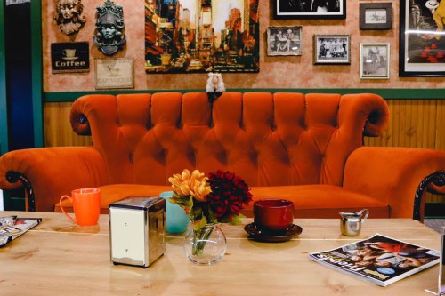 A cozy café scene featuring an orange tufted sofa, a wooden table with flowers, mugs, and a sugar container.