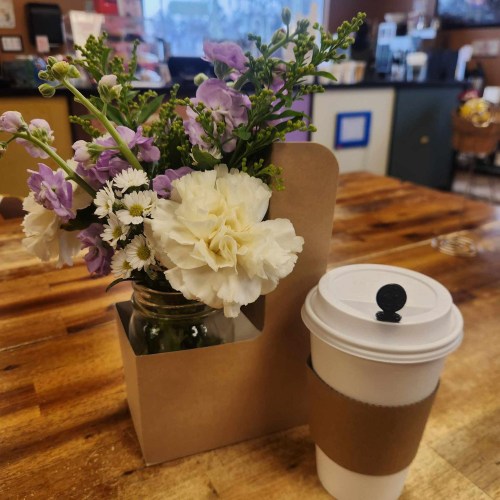 A small bouquet of flowers in a jar next to a coffee cup on a wooden table.