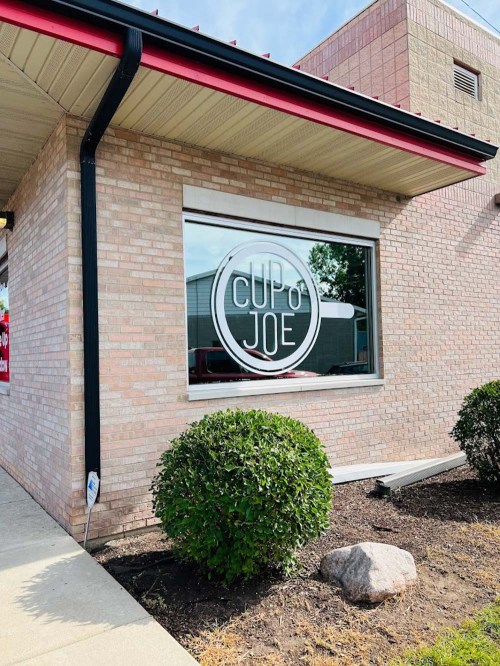 Exterior view of a brick building with a large window displaying the sign "Cup o' Joe" and landscaped bushes in front.