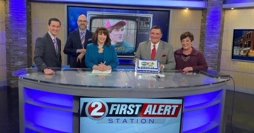 A news team stands behind a cake celebrating a milestone, with a TV screen showing a young man in the background.
