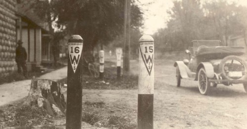 Historic street scene featuring old milestone markers and a vintage car parked nearby.