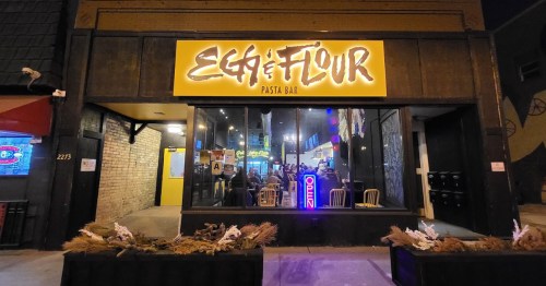 Exterior of Egg & Flour Pasta Bar, featuring a bright sign and a lively atmosphere with patrons inside.