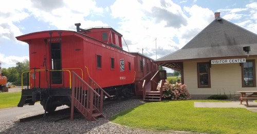 A red caboose next to a visitor center, surrounded by green grass and blooming flowers under a partly cloudy sky.