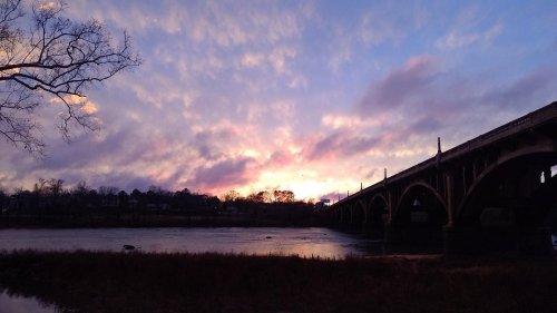 A bridge silhouetted against a colorful sunset, with clouds reflecting hues of pink and purple over a calm river.