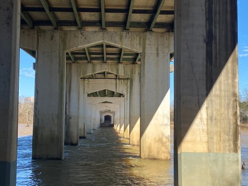 View of a bridge's understructure with concrete pillars and water flowing beneath, illuminated by sunlight.