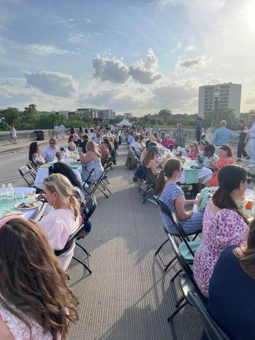 A long table set for a large outdoor dinner event, with guests enjoying food and drinks under a sunny sky.