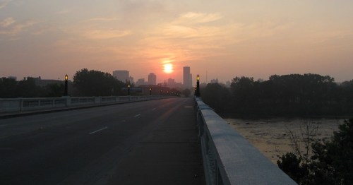 Sunset over a city skyline viewed from a bridge, with a calm river and trees in the foreground.