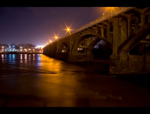 A nighttime view of a bridge over a river, illuminated by streetlights reflecting on the water's surface.