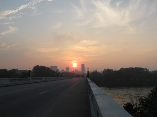 Sunset over a city skyline viewed from a bridge, with soft clouds and a calm river below.