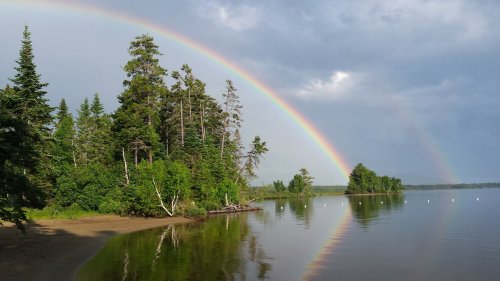 A serene lake scene with a vibrant rainbow arching over lush green trees and calm water reflecting the landscape.