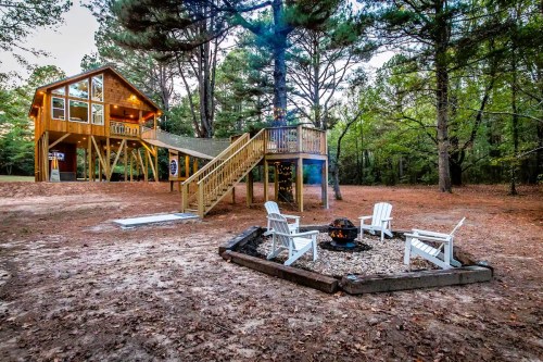A cozy cabin on stilts surrounded by trees, with a fire pit and white chairs in a serene outdoor setting.