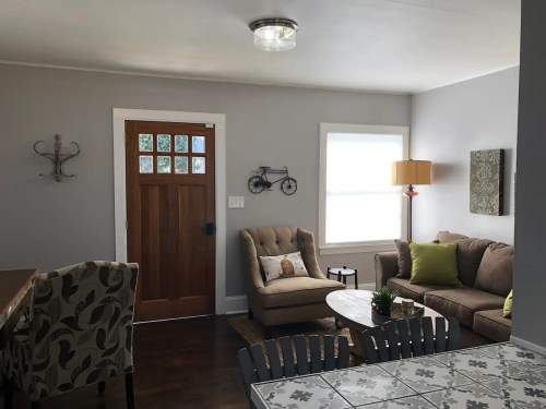 Cozy living room with a brown sofa, armchair, lamp, and a wooden door, featuring light gray walls and decorative accents.