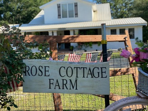 Sign for "Rose Cottage Farm" in front of a white house, with American flags and greenery in the foreground.