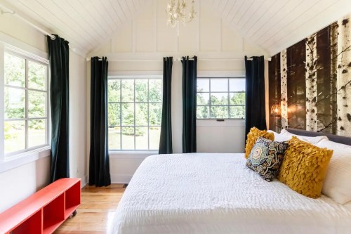 Bright bedroom with a large window, white bedding, decorative pillows, and a red shelf against a wooden accent wall.
