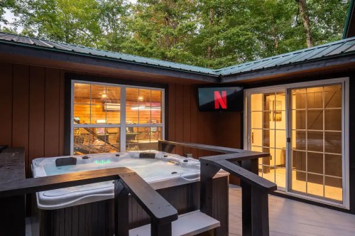 A cozy outdoor space featuring a hot tub, wooden deck, and a TV displaying the Netflix logo, surrounded by trees.